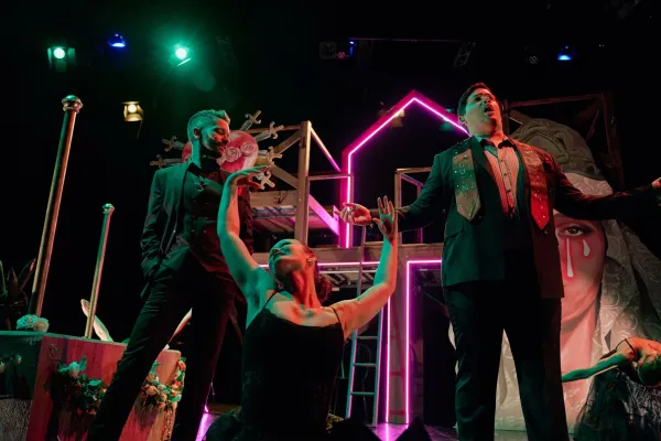 Image of 3 artists performing on a dimly lit stage with pink neon lights. One man is singing and one female appears to be dancing arms raised